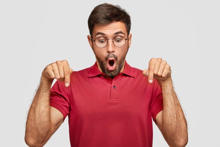 horizontal shot of stupefied young male student, looks surprisingly and indicates down, being stunned by something unbelievable, wears casual bright red t shirt, stands against white studio wall